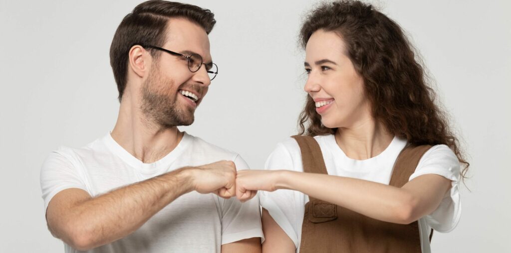 Ein lächelnder junger Mann gibt junger Frau eine Fauststoß-Pose vor grauem Hintergrund