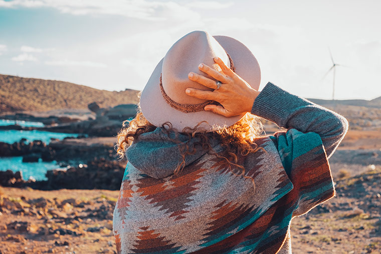 Eine Frau hält ihren Hut fest und Blickt in die fernen Dünen. Das Foto ist von hinten aufgenommen.