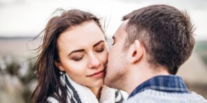 Seelenpartner in der Affäre, Mann und Frau in enger emotionaler Verbindung, sie stehen in der Natur, sie lehnt an ihn, er küsst ihre Wange, sie hat die Augen zu