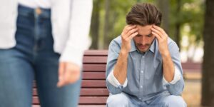 Junges Paar geschieden und Beziehung aufgelöst. Depressiver Mann sitzt auf Bank im Freien, seine Freundin verlässt