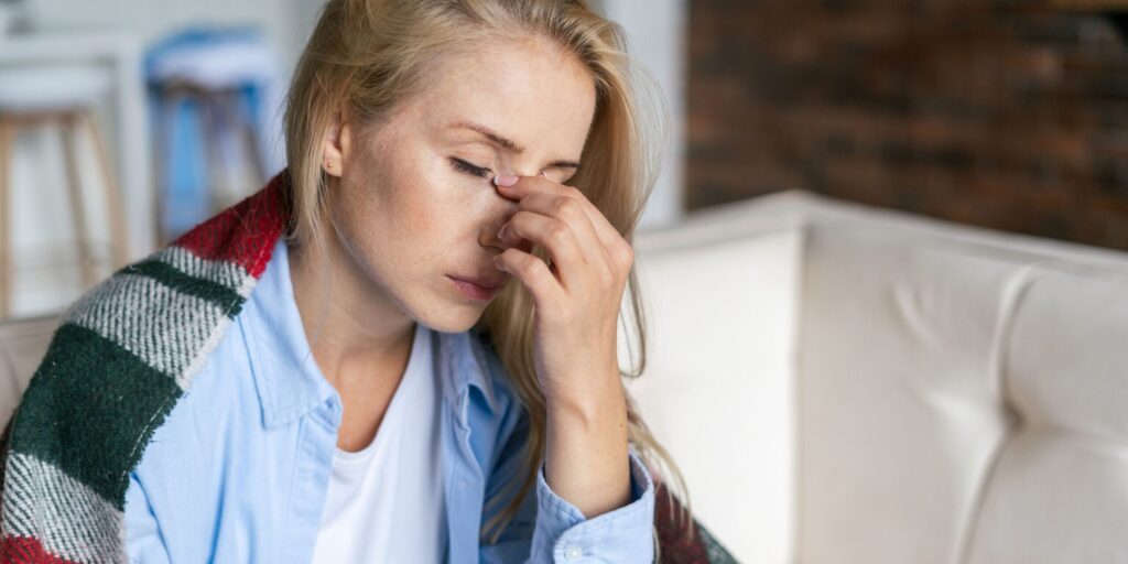 Eine müde Frau sitzt auf dem Sofa, hat die Augen geschlossen und fasst sich ins Gesicht