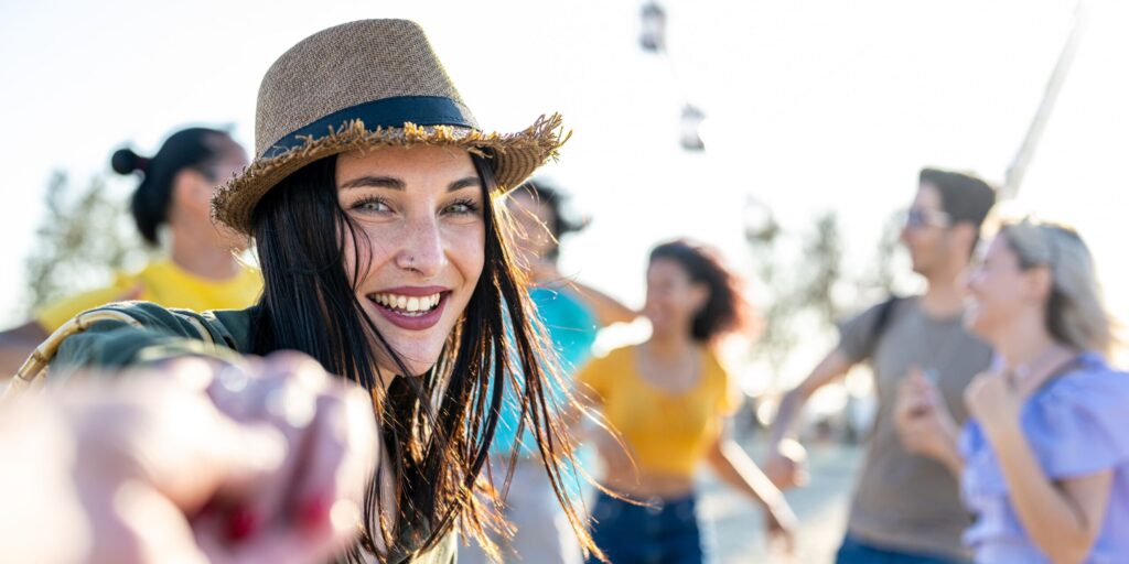 Eine junge, brünette Frau mit blauen Augen lädt dich zum Tanzen auf einer Strandparty ein, einem internationalen Musikfestival mit einer fröhlichen, großen und vielfältigen Menschenmenge.