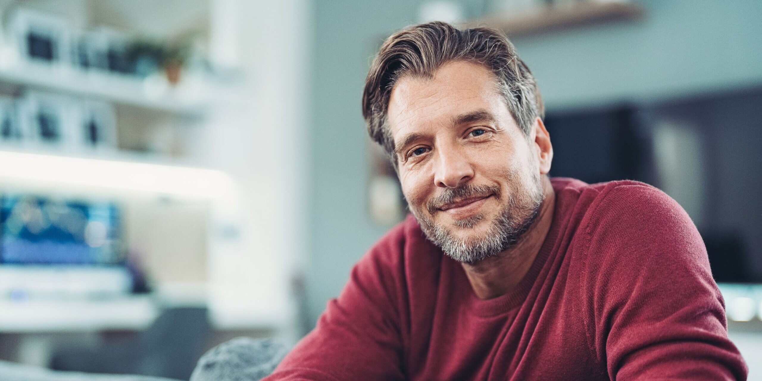 Ein MAnn mittleren alters sitzt lächelnd auf dem Sofa in seinem Wohnzimmer und schaut direkt in die Kamera.