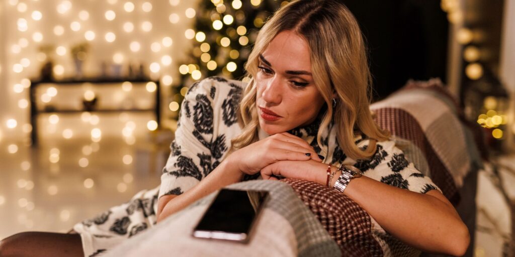 Porträt einer jungen blonden Frau, die an der Rückenlehne des Sofas lehnt, auf ihr Smartphone schaut und auf den Empfang einer SMS von einem geliebten Menschen wartet.