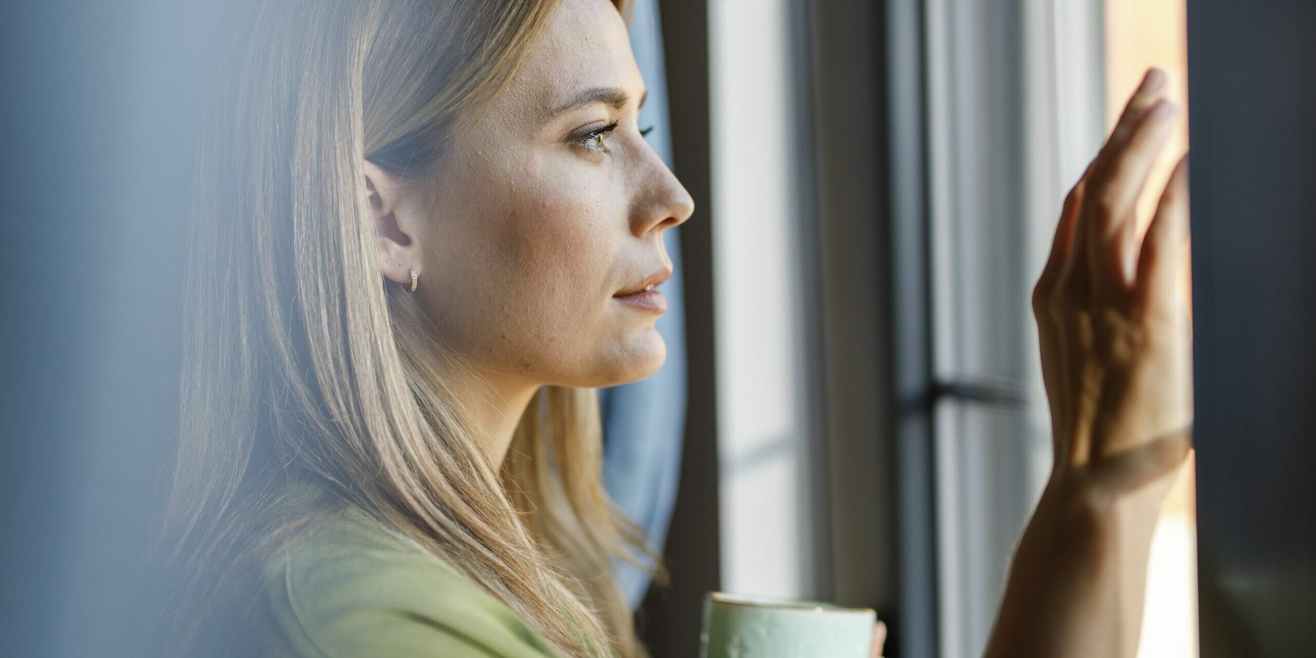 Porträt einer schönen Frau mittleren Alters, die am Fenster steht, nach draußen schaut, lächelt und nachdenkt, während sie eine Tasse Morgenkaffee genießt.
