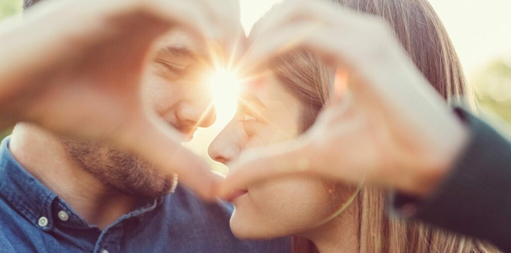 Frau und Mann machen das Herz-Symbol per Hand, hinter ihnen ein Sonnenuntergang