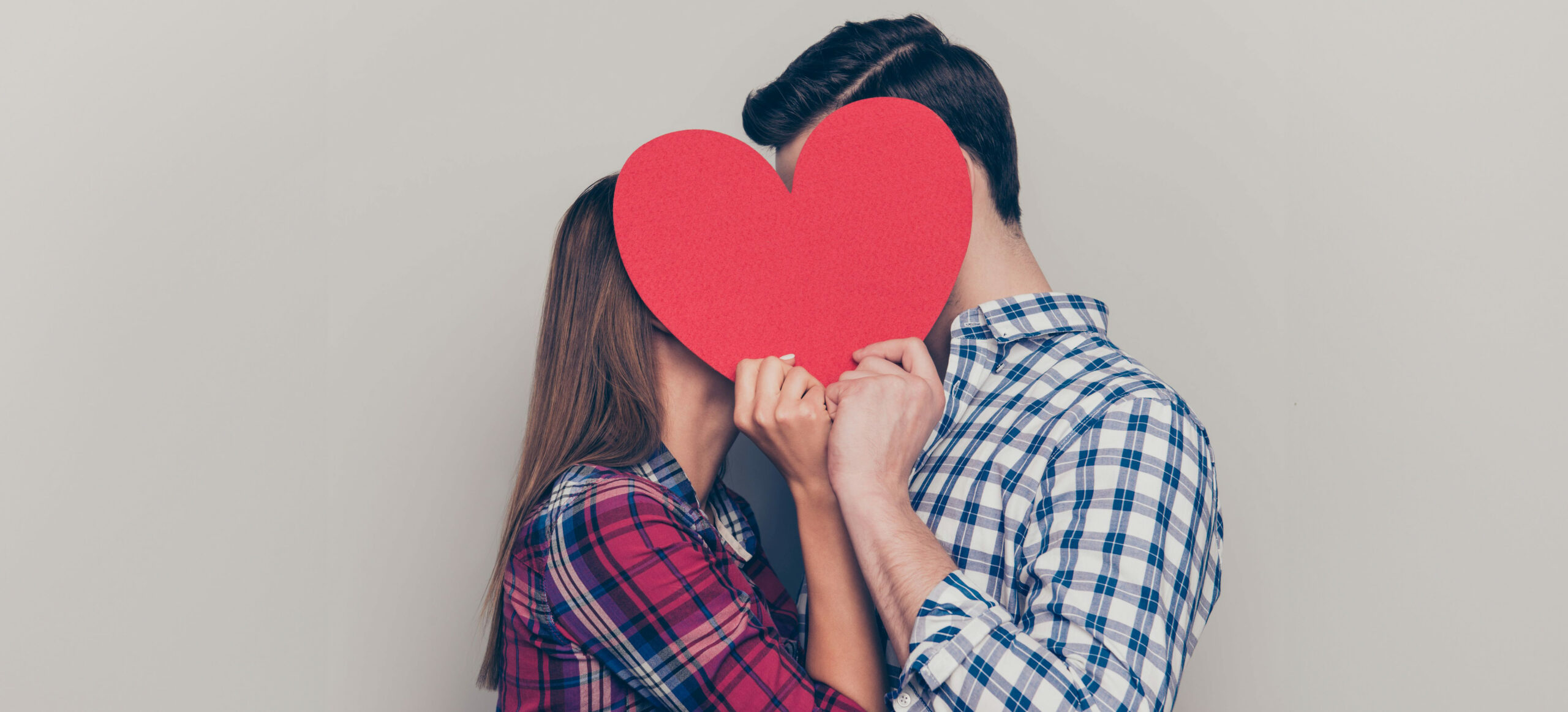 Ein Paar mit karierten Hemden küsst sich hinter einem roten Herz aus Papier und grauem Hintergrund.
